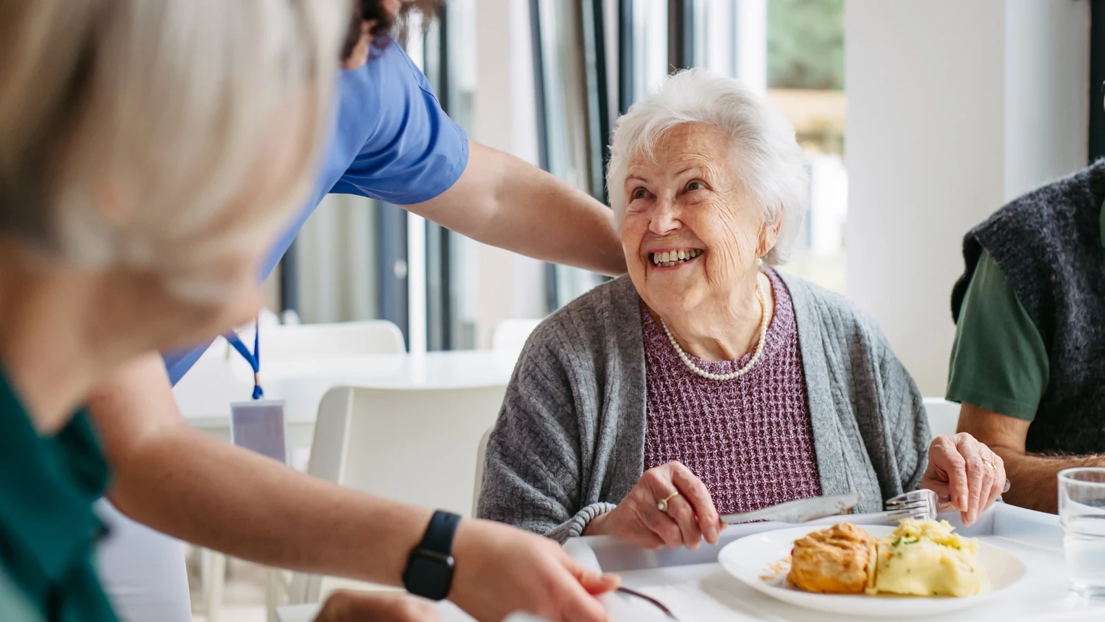 Mujer mayor sonriendo mientras una cuidadora le sirve el almuerzo